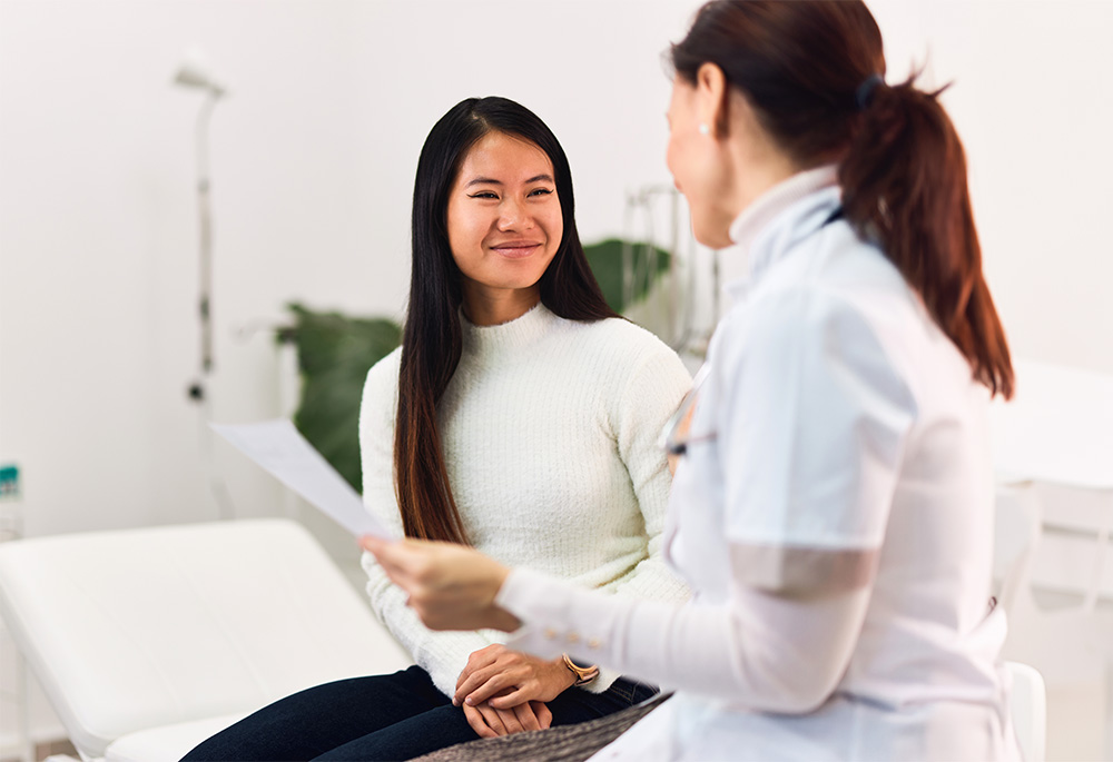 A smiling Asian female patient talking with a female doctor, sitting in the medical office.